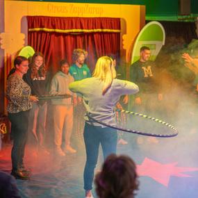 Eine Frau mit Hula-Hoop-Reifen steht im Fokus einer Zirkusauff&uuml;hrung, umgeben von Zuschauern und Nebel.