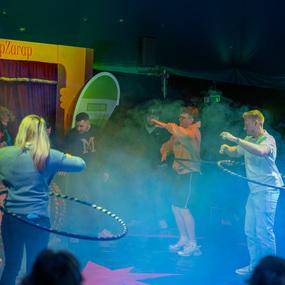 F&uuml;nf Personen mit Hula-Hoops tanzen in einem rauchigen Zelt w&auml;hrend einer bunten Veranstaltung.