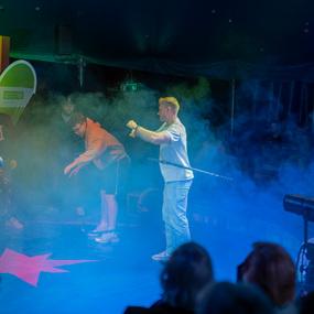 Leute stehen in einem Zirkuszelt, umgeben von Nebel und bunten Lichtern, w&auml;hrend sie verschiedene akrobatische Tricks versuchen.