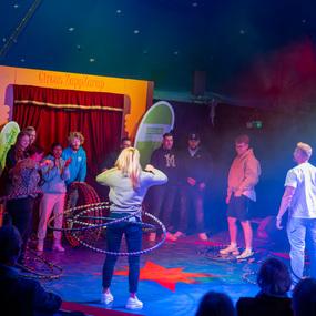 Zuschauer im Zirkuszelt beobachten eine Hula-Hoop-Performerin vor einem bunten Hintergrund.