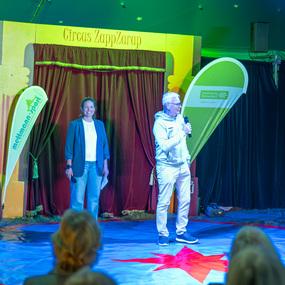 Zwei Personen stehen auf einer Zirkusbühne, mit buntem Hintergrund und einem roten Stern auf dem Boden.