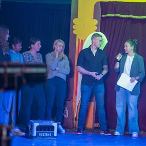 Gruppe von sechs Personen auf einer B&uuml;hne; eine Frau mit Mikrofon spricht, w&auml;hrend die anderen aufmerksam zuh&ouml;ren.