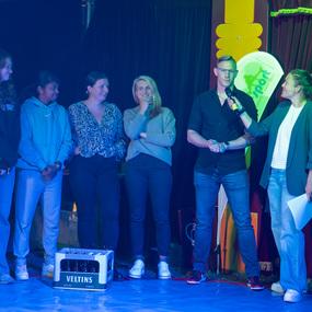 Sechs Personen stehen auf einer B&uuml;hne, w&auml;hrend eine Frau mit Mikrofon einen Mann in schwarzem Hemd interviewt.