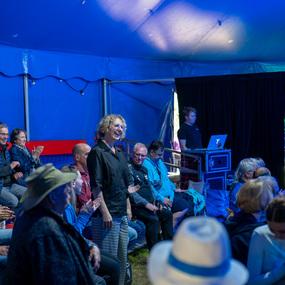 Menschen sitzen in einem Zelt und applaudieren, während eine Frau in der Mitte lächelt. Im Hintergrund steht ein DJ.