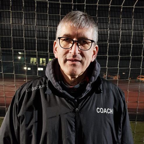Mann mit Brille und grauen Haaren steht im Dunkeln mit einem „Coach“-Wortlaut auf seiner Jacke vor einem Fußballtor.