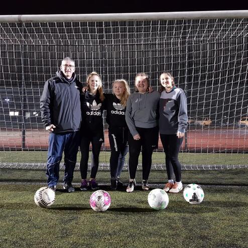 Gruppe aus fünf Personen steht vor einem Fußballtor, mit drei Fußbällen zu ihren Füßen auf einem Kunstrasenplatz.