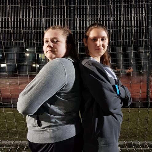 Zwei Jugendliche stehen Rücken an Rücken vor einem Sportplatz, beleuchtet von Stadtlichtern im Hintergrund.