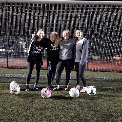 Vier Mädchen stehen lächelnd vor einem Fußballtor, zwei Fußbälle liegen auf dem Kunstrasen.