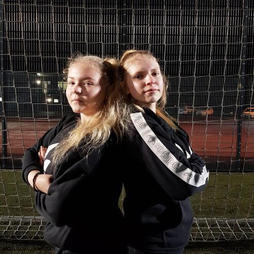 Zwei Mädchen stehen Rücken an Rücken vor einem Fußballtor und posieren in schwarzen Sweatshirts.