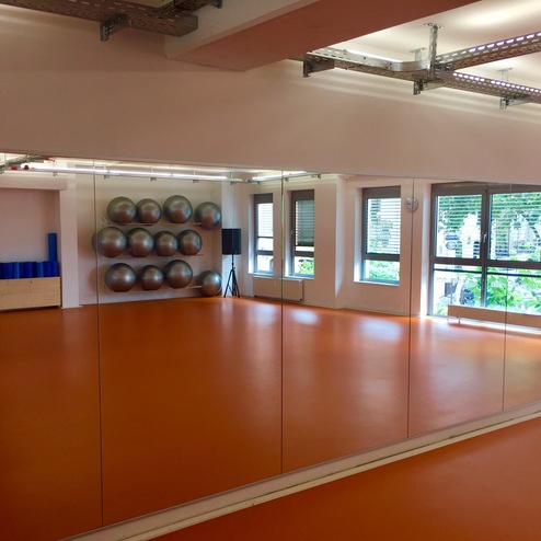 Fitnessraum mit Spiegelwand, statischen Übungen, orangefarbenem Boden und einer Wand mit mehreren Fitnessbällen.