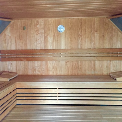 Innenansicht einer Holzsauna mit Bänken, Thermometer und abgeschrägten Ecken. Wände sind aus hellem Holz.