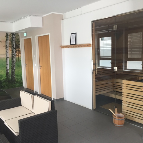 Wohlfühlbereich mit Sitzgelegenheiten, Zugang zur Sauna und birkenbedeckter Wandgestaltung. Holzfass mit Schöpfkelle im Vordergrund.