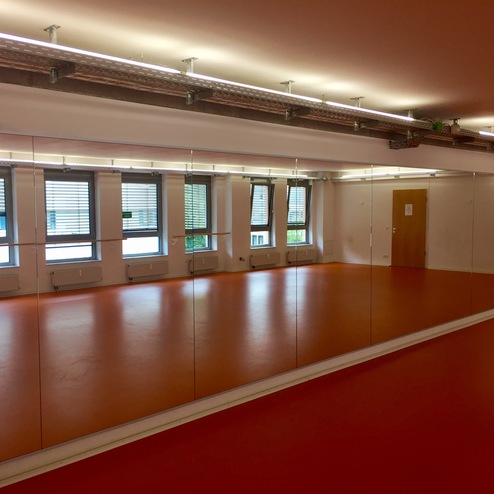 Spiegelwand in einem Raum mit orangefarbenem Boden und großen Fenstern für Tageslicht.