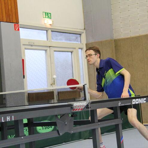 Ein Tischtennisspieler in blauer Sportkleidung schlägt mit einem Schläger einen Ball über das Netz auf einem Tischtennistisch.