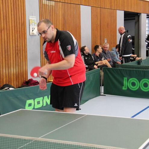 Spieler in rotem Shirt serviert einen Tischtennisball an einem grünen Tischtennistisch, Zuschauer im Hintergrund.