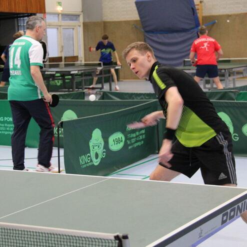 Tischtennisspieler in Aktion, während er einen Ball über ein Netz auf einem grünen Tisch sendet.