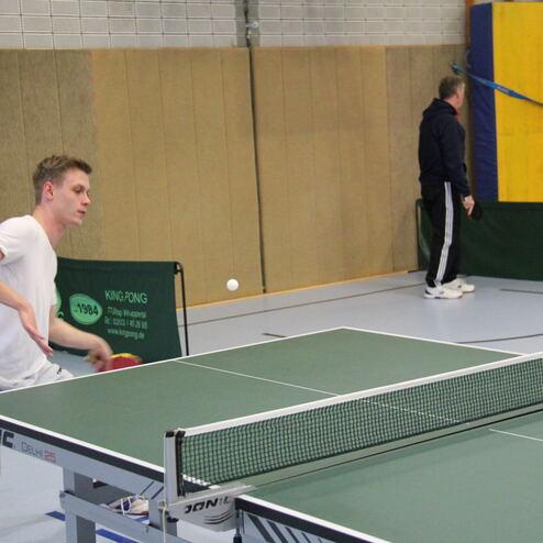 Junger Mann schlägt mit einem Tischtennisschläger einen Ball über das Netz auf einer Tischtennisplatte.