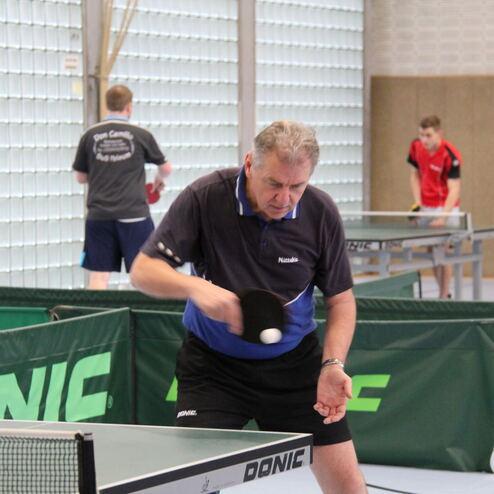 Ein Mann spielt Tischtennis, konzentriert auf den Ball, während zwei weitere Spieler im Hintergrund aktiv sind.