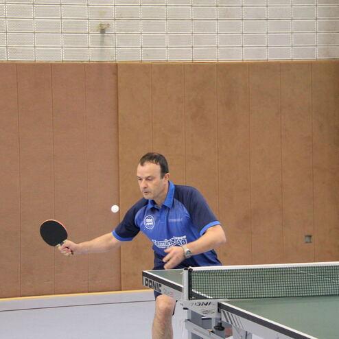Tischtennisspieler in blauer Sportkleidung schlägt einen Ball auf einem Tischtennistisch. Hintergrund mit Holzpaneelen.