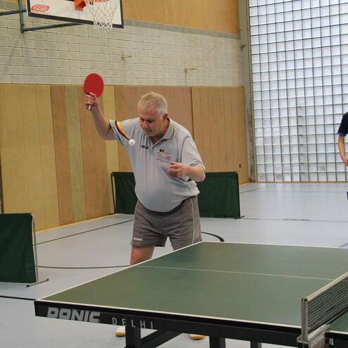 Ein älterer Mann spielt Tischtennis und hebt den Schläger, während ein Jugendlicher am Tisch steht.