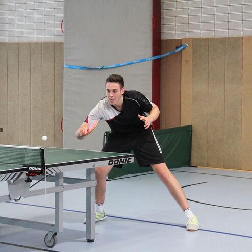 Junger Mann spielt Tischtennis, bereit zum Schlag, während der Ball über das Netz fliegt.