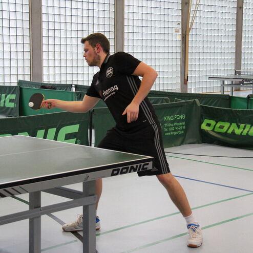 Spieler schlägt mit einem Tischtennisschläger einen Ball auf einer Tischtennisplatte in einer Sporthalle.