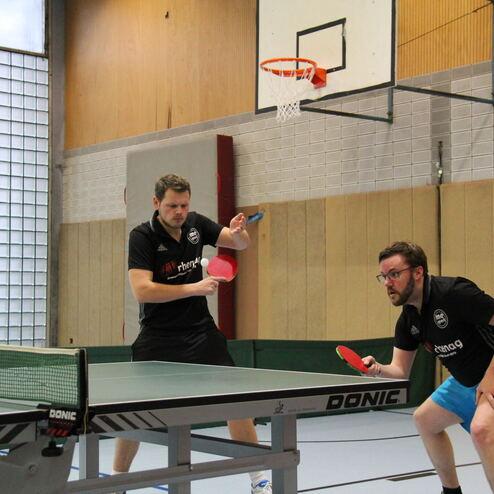 Zwei Männer spielen Tischtennis in einer Halle, einer bereit für den Aufschlag, der andere in Erwartung.
