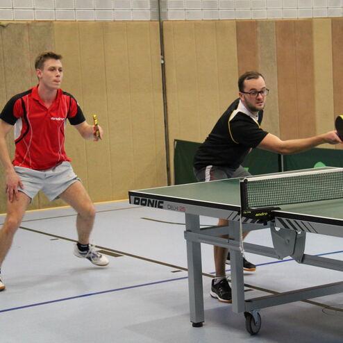 Zwei Tischtennisspieler in Aktion, einer serviert, der andere bereitet sich auf den Schlag an einem Tischtennistisch vor.