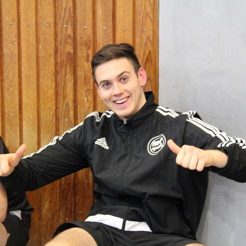 Zwei junge Männer sitzen an einer Wand; einer lächelt und zeigt Daumen hoch, der andere schaut lachend auf ihn.