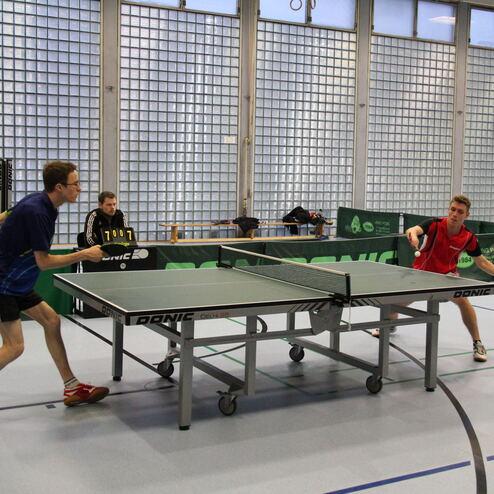 Zwei Tischtennisspieler im Wettkampf, während ein Schiedsrichter die Punkte zählt. Tischtennisplatten im Hintergrund.