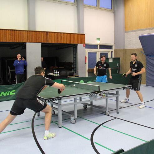 Vier Tischtennisspieler in einem Wettkampf an einem Tischtennistisch in einer Sporthalle.