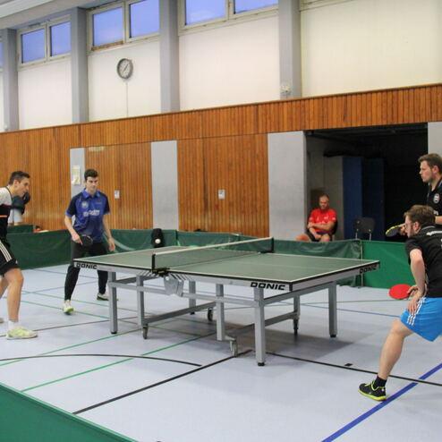 Zwei Tischtennisspieler stehen an einem Tisch, während ein dritter Spieler bereit ist, zu schlagen. Zuschauer im Hintergrund.