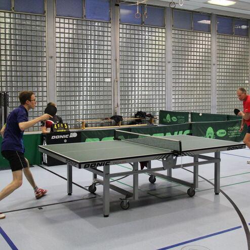 Zwei Tischtennisspieler in Aktion an einem Tischtennistisch in einer Halle mit grünen Wänden und Fenstern.