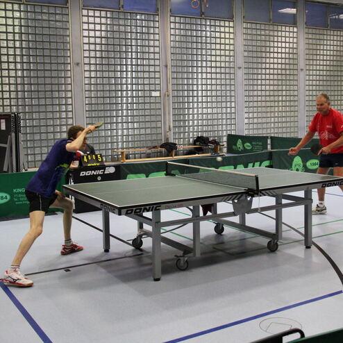 Zwei Tischtennisspieler in einem Sportraum spielen auf einer grünen Tischtennisplatte. Im Hintergrund ist ein Punktestand sichtbar.