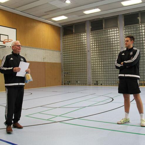 Zwei Personen in Sportbekleidung stehen in einer Sporthalle. Der ältere Mann hält einen Zettel und spricht.