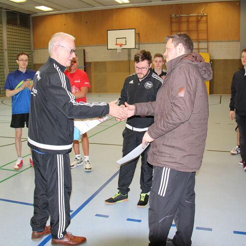 Zwei Männer schütteln sich die Hände, während andere Personen applaudieren. Sporthalle mit Tischtennisplatte im Hintergrund.