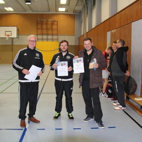 Drei Männer in Sportbekleidung halten Urkunden, umgeben von weiteren Teilnehmern in einer Sporthalle.