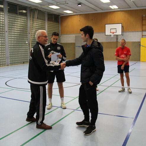 Zwei Männer schütteln sich die Hände in einer Sporthalle, während zwei weitere Personen im Hintergrund zuschauen.