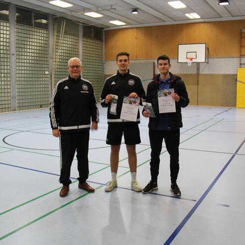 Drei Personen in Sportkleidung stehen in einer Halle und halten Urkunden in den Händen. Basketballkorb im Hintergrund.