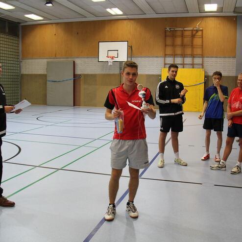 Junger Tischtennisspieler hält einen Pokal, während Trainer und andere Spieler in einer Sporthalle zuschauen.