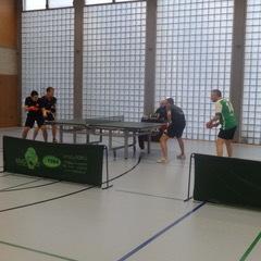 Zwei Tischtennisspieler stehen an einem Tisch, während zwei Gegner sich zum Schlag vorbereiten. Sporthalle mit Fensterlicht.