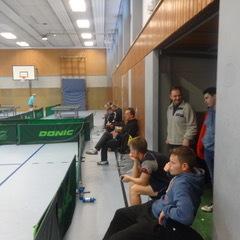 Gruppierung von Personen am Rand einer Tischtennishalle, während im Hintergrund ein Spiel stattfindet.