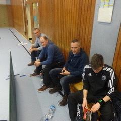 Vier Männer sitzen auf einer Bank in einer Sporthalle, die Wände sind aus Holz und der Boden blau.