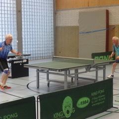 Zwei ältere Männer spielen Tischtennis an einem Tisch in einer Sporthalle.