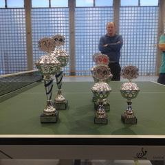 Trophäen stehen auf einem Tischtennistisch, im Hintergrund zwei Männer beobachten die Szenarie.