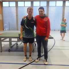 Zwei junge Männer stehen lächelnd arm in arm vor einem Tischtennistisch in einer Sporthalle.