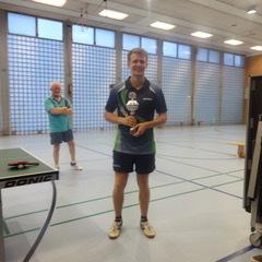 Ein Spieler hält einen Pokal in einer Sporthalle, während andere Zuschauer im Hintergrund stehen.