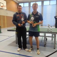 Zwei Tischtennisspieler halten Pokale in einer Sporthalle, im Hintergrund eine Tischtennisplatte und weitere Personen.