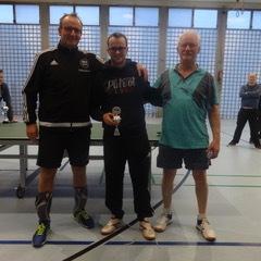 Drei Männer posieren nach einem Tischtennisturnier, einer hält einen Pokal in der Mitte, mit Tischtennisplatte im Hintergrund.