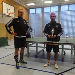 Zwei Sportler in Trikots stehen neben einem Tischtennistisch, einer hält einen Pokal in den Händen.